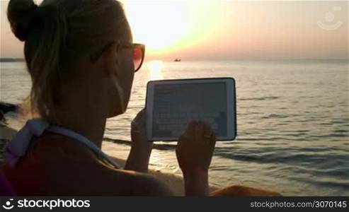 Woman typing on tablet computer sitting on beach at sunset. Vacation with pad