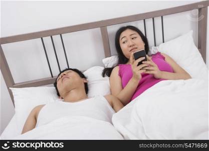 Woman using cell phone while looking at man sleeping in bed