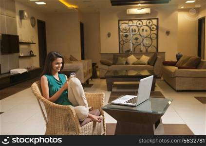 Woman using cell phone while sitting on wicker chair in front of coffee table with laptop at home