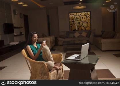 Woman using cell phone while sitting on wicker chair in front of coffee table with laptop at home