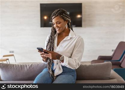Woman using cellphone in her home and sitting on the back of the couch. African American woman with braids inside of home using cellphone.. Woman using cellphone in her home and sitting on the back of the couch