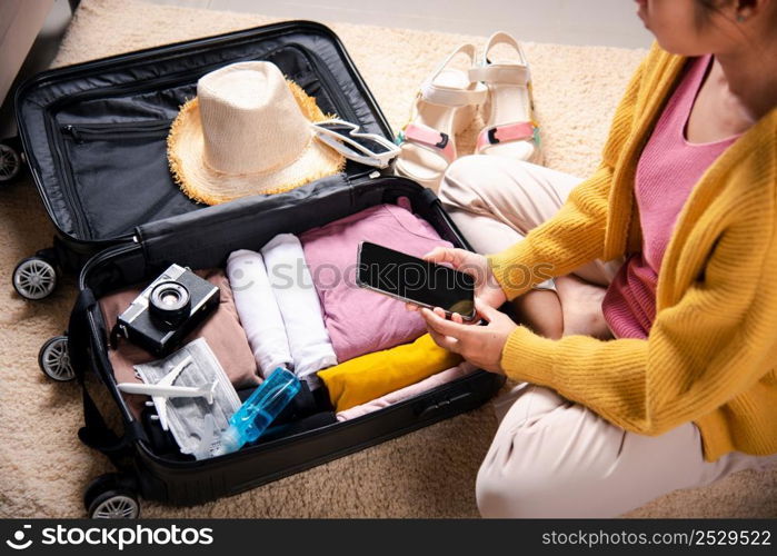 Woman using smartphone blank screen and packing suitcase bag for travel trip weekend vacation at home, female check information in tourism website on mobile phone before go to airport