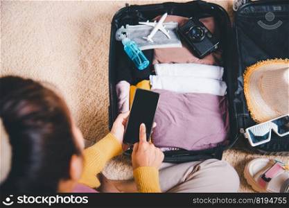 Woman using smartphone blank screen and packing suitcase bag for travel trip weekend vacation at home, female check information in tourism website on mobile phone before go to airport