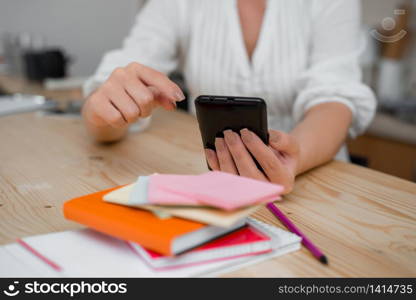 woman using smartphone office supplies technological devices inside home. Young lady using a dark smartphone inside home. Woman holding a black cell phone in a house ambient. Girl with technological devices and office supplies with blank space.