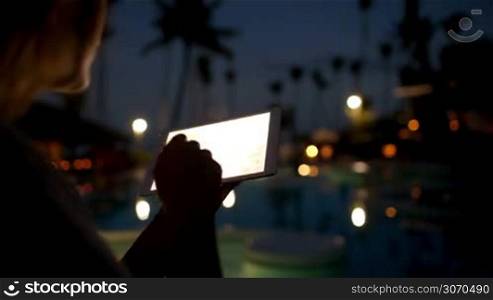 Woman using tablet computer on tropical resort in the evening. Pad screen shining brightly