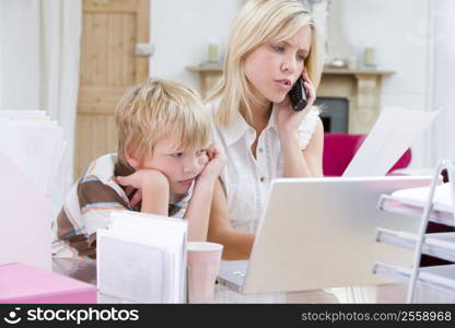 Woman using telephone in home office with laptop while young boy waits