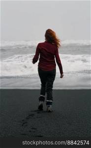 Woman walking along Reynisfjara beach scenic photography. Picture of person with rough sea on background. High quality wallpaper. Photo concept for ads, travel blog, magazine, article. Woman walking along Reynisfjara beach scenic photography