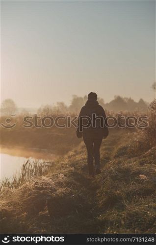 Woman walking through a meadow by a pond in the foggy morning. Sun rising above field and pond flooded with fog in the morning. Real people, authentic situations