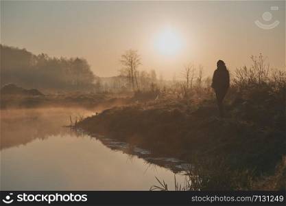 Woman walking through a meadow by a pond in the foggy morning. Sun rising above field and pond flooded with fog in the morning. Real people, authentic situations