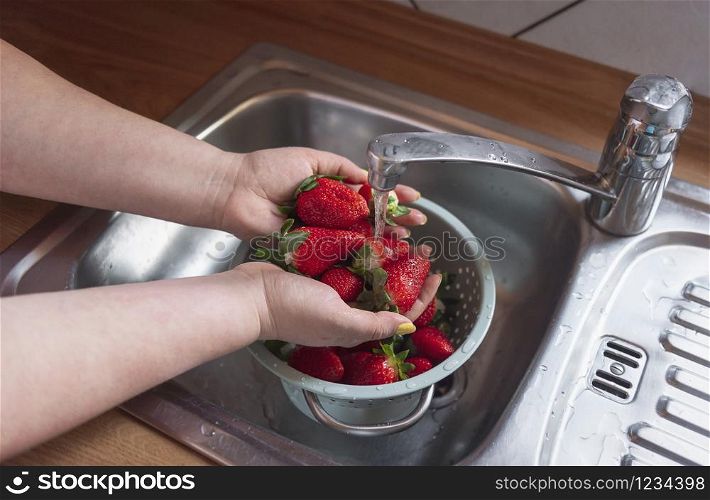 Woman washing ripe strawberries in the kitchen sink, under tap water string. Organic strawberry fruits in colander. Sweet summer fruits. Home cooking