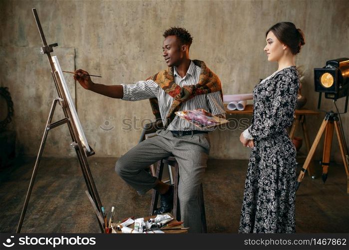 Woman watches as painter with a palette draws with a brush. Man sitting at the easel, art studio interior on background, creative master in workshop. Woman watches as painter draws with a brush