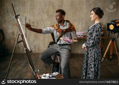 Woman watches as painter with a palette draws with a brush. Man sitting at the easel, art studio interior on background, creative master in workshop. Woman watches as painter draws with a brush