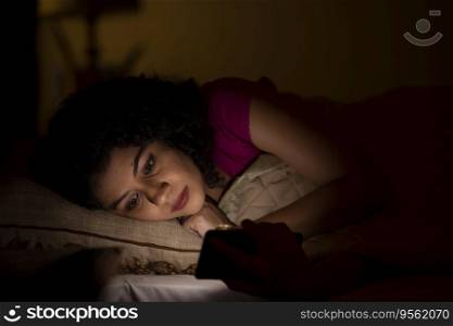 Woman watching smartphone alone at night while lying on bed 
