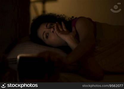 Woman watching smartphone with sleepy eyes at night while lying on bed 