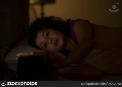 Woman watching smartphone with sleepy eyes at night while lying on bed 