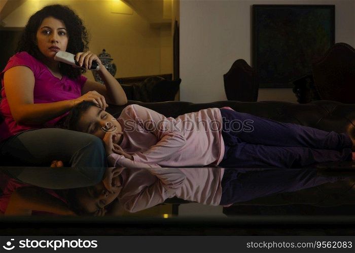 Woman watching television while her daughter sleeping on her lap on sofa 