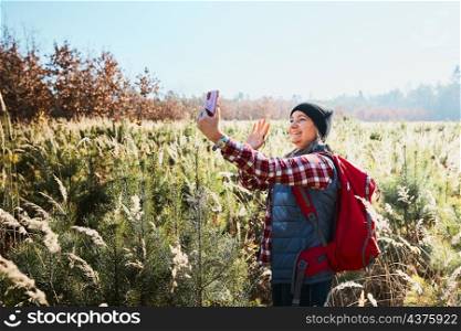 Woman waving during video call sending greetings from vacation trip in mountains. Woman with backpack hiking through tall grass along path on meadow. Spending summer vacation close to nature