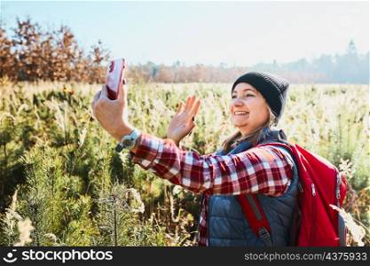 Woman waving during video call sending greetings from vacation trip in mountains. Woman with backpack hiking through tall grass along path on meadow. Spending summer vacation close to nature