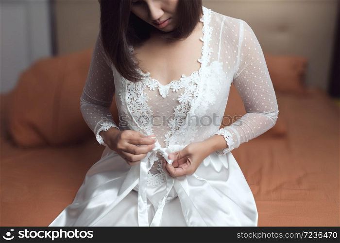 Woman wearing a long white nightgown and bow tie on a white silk robe with lace sleeve in the bedroom at night. Asian woman trying on new white nightwear for sleep