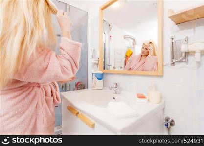 Woman wearing dressing gown brushing her long blonde hair, morning beauty routine. Haircare and hairstyling concept.. Woman wearing dressing gown brushing her hair