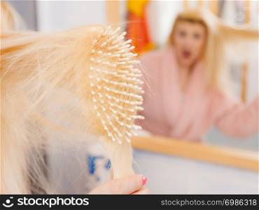 Woman wearing dressing gown trying to brush her long blonde very tangled hair, morning beauty routine. Haircare problem and hairstyling concept.. Shocked woman wearing dressing gown brushing her hair