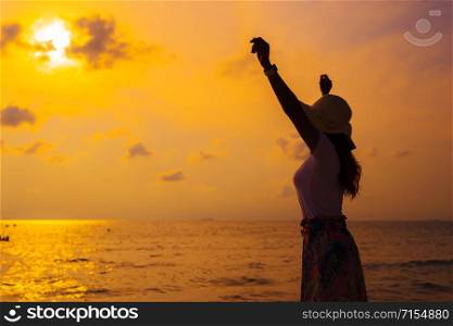 woman wearing hat with arms raised standing on the sea beach at sunset