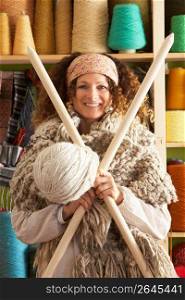 Woman Wearing Knitted Scarf Standing In Front Of Yarn Display Holding Giant Needles And Ball Of Wool