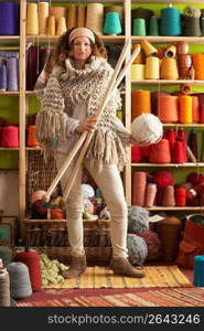 Woman Wearing Knitted Scarf Standing In Front Of Yarn Display Holding Giant Needles