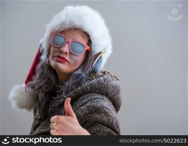 Woman wearing Santa Claus hat and sunglasses listening to music with her headphones and thumbs up. Winter season series fashion portrait