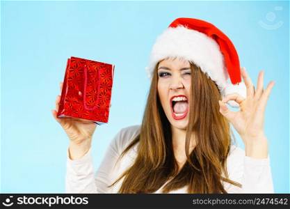 Woman wearing santa claus hat holds red gift bag, excited face expression, showing okay hand sign. Christmes time.. Excited christmas woman holds gift bag
