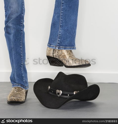 Woman wearing snakeskin cowboy boots holding foot as if to stomp on black cowboy hat.