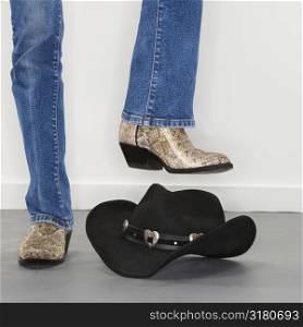 Woman wearing snakeskin cowboy boots holding foot as if to stomp on black cowboy hat.