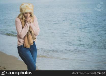 Woman wearing warm stylish autumnal outfit with furry big hat yet feeling cold. Female having chills while relaxing walking on beach.. Woman in stylish outfit feeling cold by the sea