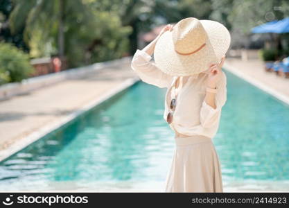 Woman wearing white shirt, long skirt and straw hat posing near swimming pool.