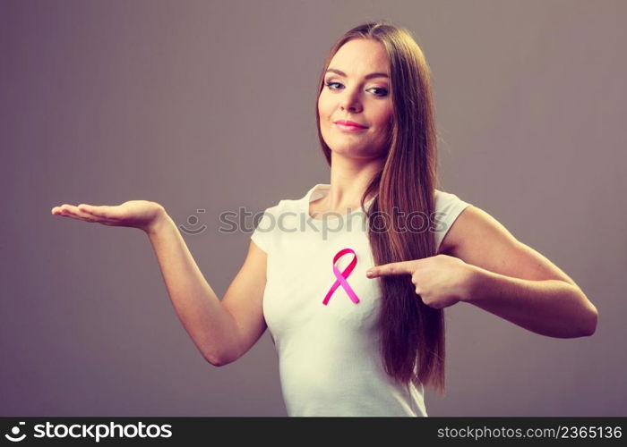 Woman wih pink cancer ribbon on chest holding open hand for product text, pointing with finger. Health care, medicine and breast cancer awareness concept. Studio shot on gray. Woman pink cancer ribbon on chest holds open hand