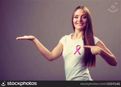 Woman wih pink cancer ribbon on chest holding open hand for product text, pointing with finger. Health care, medicine and breast cancer awareness concept. Studio shot on gray. Woman pink cancer ribbon on chest holds open hand