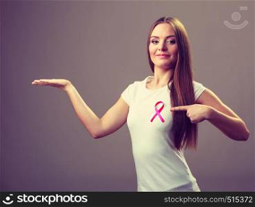 Woman wih pink cancer ribbon on chest holding open hand for product text, pointing with finger. Health care, medicine and breast cancer awareness concept. Studio shot on gray. Woman pink cancer ribbon on chest holds open hand