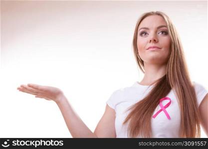 Woman wih pink cancer ribbon on chest holding open hand for product text. Health care, medicine and breast cancer awareness concept. Studio shot on gray. Woman pink cancer ribbon on chest holds open hand