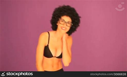 Woman with a black afro hairdo wearing black underwear laughing and waving at the camera against a purple studio background