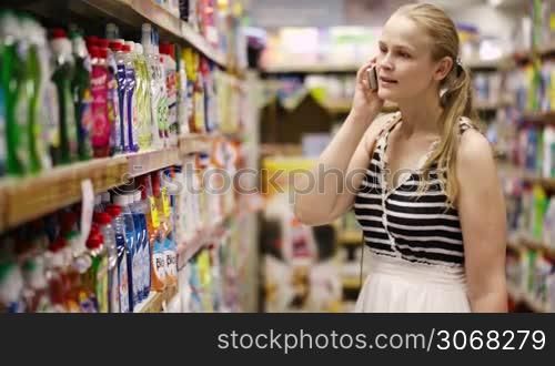 Woman with a long blond ponytail chatting on her mobile while out shopping standing in an aisle in the supermarket