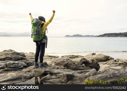 Woman with backpack and arms raised enjoying the beautiful morning view of the coast