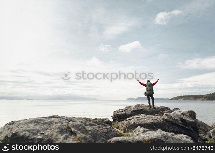Woman with backpack and arms raised  enjoying the beautiful view of the coast