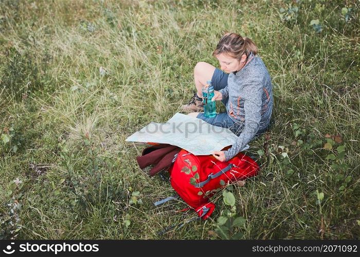 Woman with backpack having break during trip in mountains looking at map sitting on grass on summer vacation day