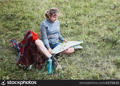 Woman with backpack having break during trip in mountains looking at map sitting on grass on summer vacation day