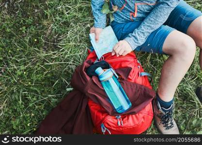 Woman with backpack having break during trip in mountains taking map out sitting on grass on summer vacation day