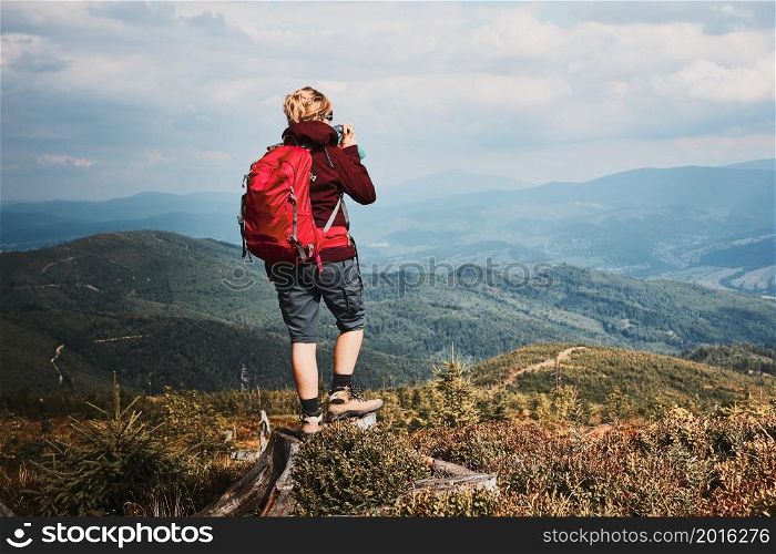 Woman with backpack hiking in mountains. Spending summer vacation close to nature. Rear view of woman standing on stump on top of hill admiring mountain landscape panorama