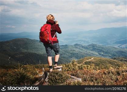 Woman with backpack hiking in mountains. Spending summer vacation close to nature. Rear view of woman standing on stump on top of hill admiring mountain landscape panorama