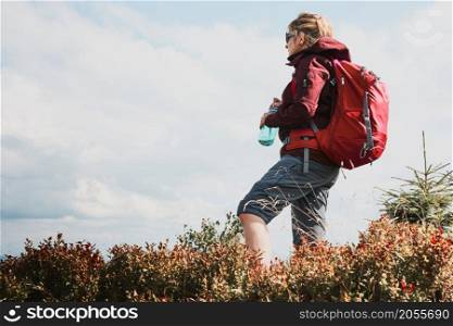 Woman with backpack hiking in mountains, spending summer vacation close to nature. Woman standing on top of hill admiring mountain landscape panorama holding bottle with water