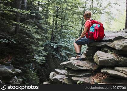 Woman with backpack hiking in mountains. Woman taking break sitting on rock. Spending summer vacation close to nature