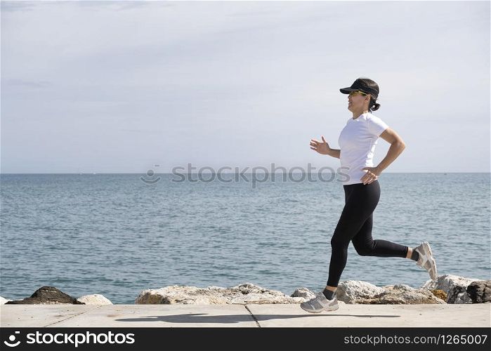 woman with cap and sunglasses running along the promenade, the sea in the background